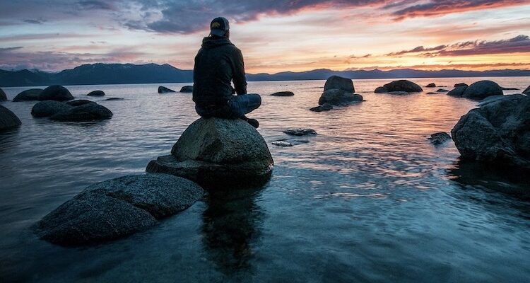 Photo credit: Keegan Houser [Person sitting on rock near shoreline meditating during sunset]