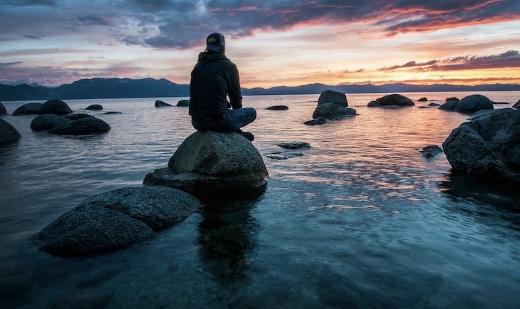 Photo credit: Keegan Houser [Person sitting on rock near shoreline meditating during sunset]