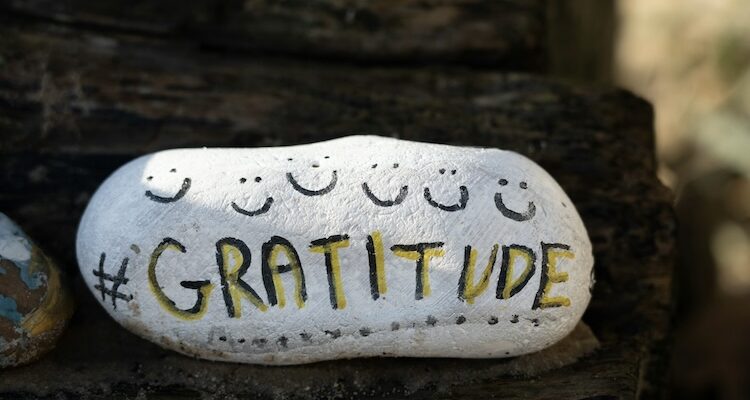 White painted rock with six smiley faces and the word "#GRATITUDE" in black and yellow letters, a reminder to practice gratitude in sports
