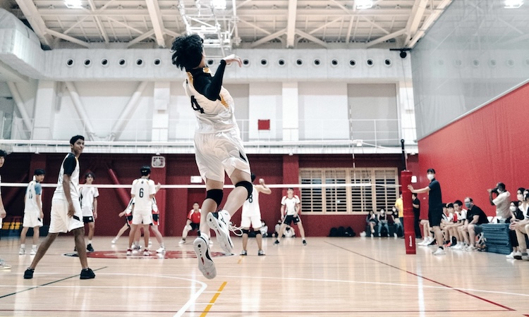 Volleyball player leaping to hit a jump serve in an indoor gym as teammates wait on the court