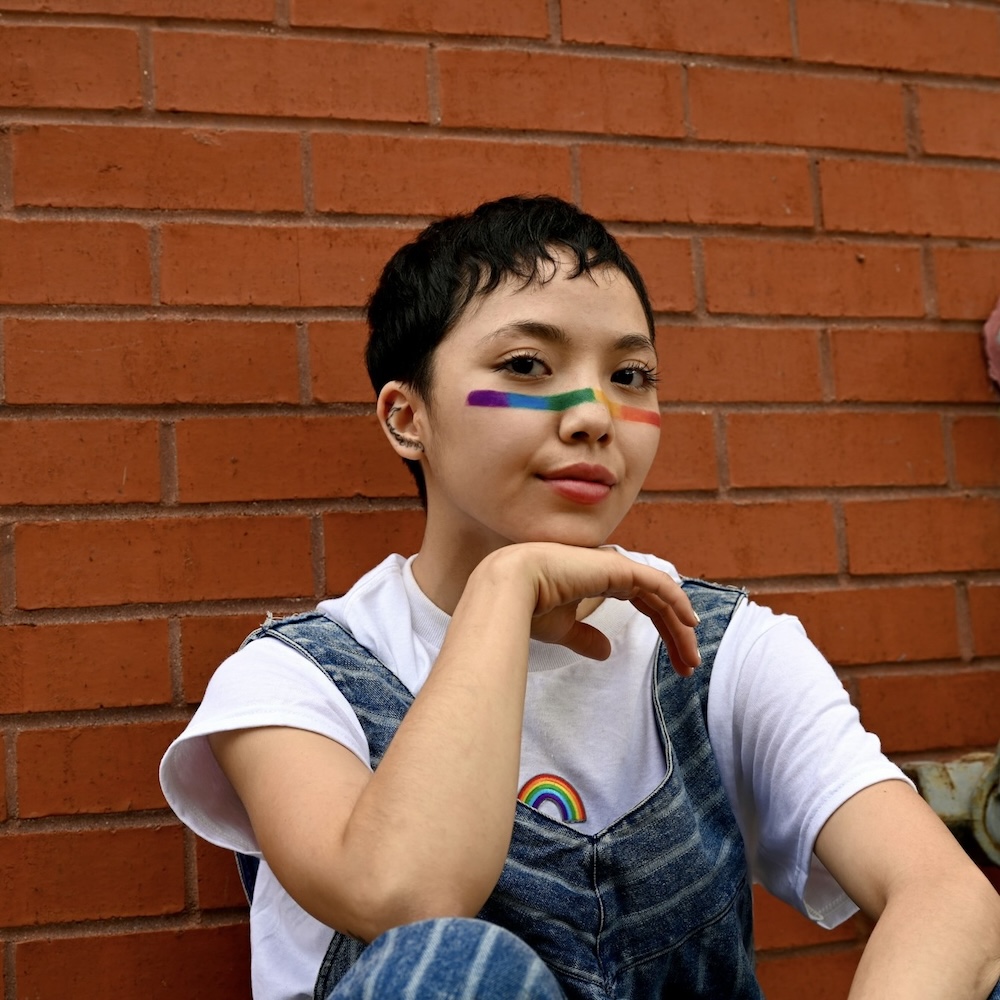 a person leaning against a brick wall wearing a white shirt and jean overalls with a rainbow patch and a rainbow painted face depictive of queer and gender neutral identity who might benefit from LGBTQ+ Counseling Colorado.