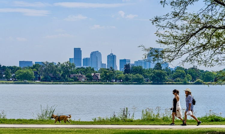 Two people and a dog walk along a lakeside path with the downtown Denver skyline in the background